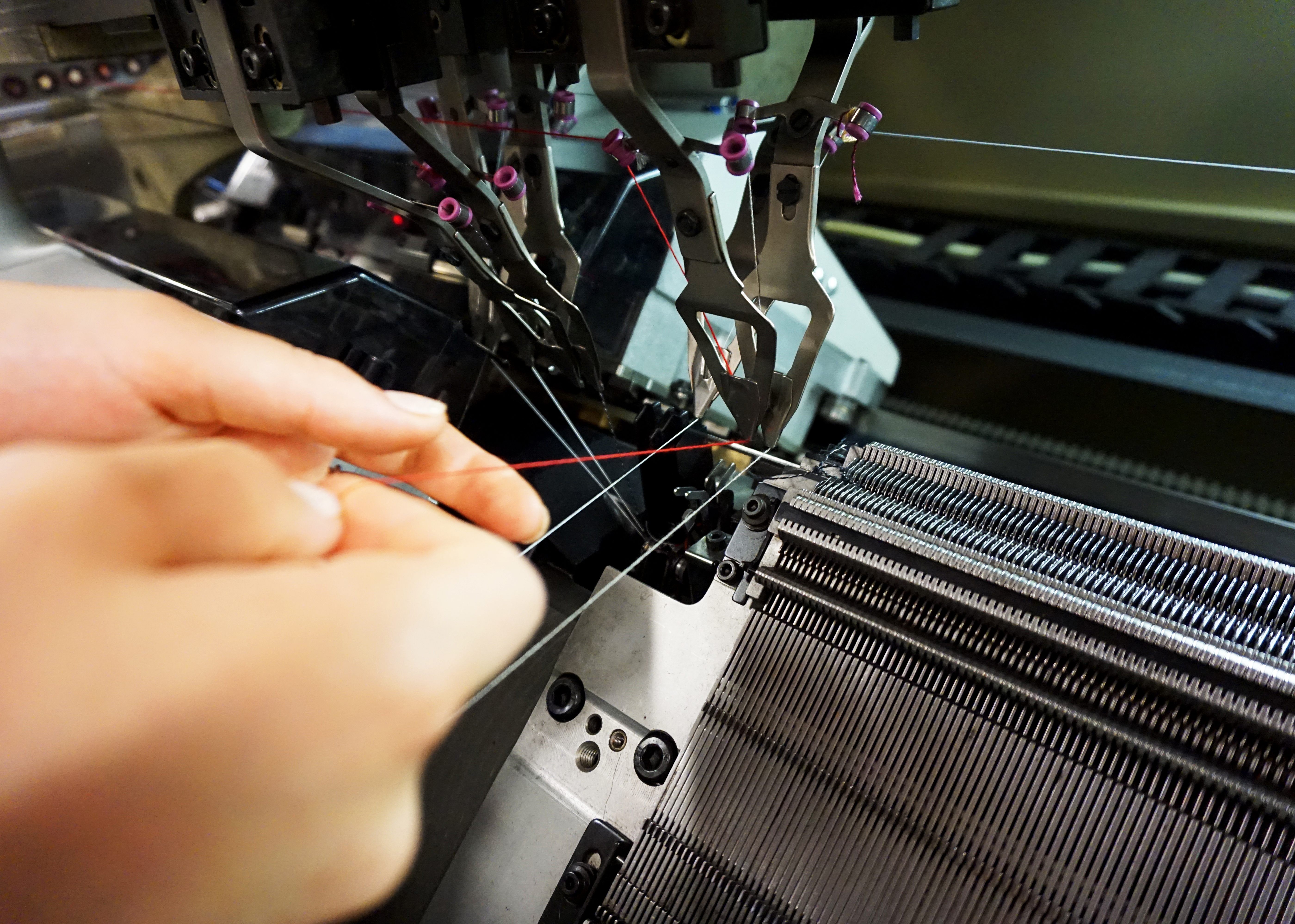 A close-up photo of a person's hands guiding threads in an industrial knitting machine